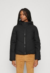 Veste matelassée noire avec capuche, dotée de coutures en chevron, fermeture éclair à l'avant et poignets élastiques. Portée sur un pantalon en velours côtelé marron clair.