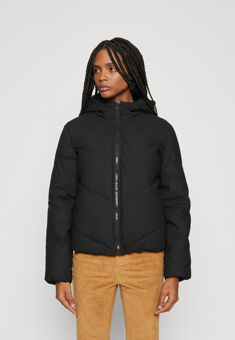 Veste matelassée noire avec capuche, dotée de coutures en chevron, fermeture éclair à l'avant et poignets élastiques. Portée sur un pantalon en velours côtelé marron clair.