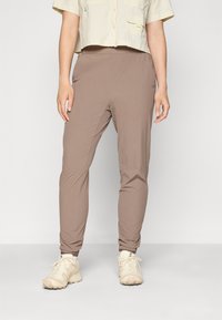 Bruine, taps toelopende broek van lichtgewicht stof met een gladde textuur. Heeft een elastische tailleband en aansluitende boorden, gecombineerd met lichte schoenen.