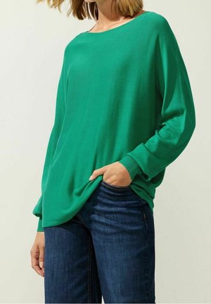 Kvinde iført en løs grøn langærmet sweater med ribbede manchetter og mørkeblå jeans, den ene hånd i lommen.