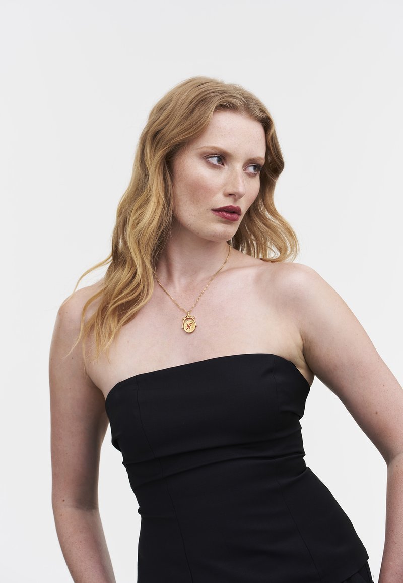 Zwarte strapless top met een nauwsluitend ontwerp. Opvallende gouden ketting met een ronde hanger. Model heeft golvend haar en een neutrale achtergrond.