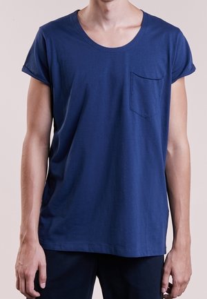 Homme portant un t-shirt bleu marine uni à manches courtes avec une petite poche sur la poitrine, se tenant devant un fond neutre.