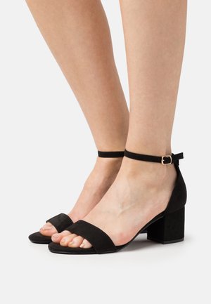 Pieds portant des sandales en daim noir à talons blocs avec brides à la cheville et orteils découverts sur un fond clair uni.