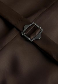 Brauner Stoff mit glatter Textur, versehen mit einer silbernen Metallschnalle. Die Nähte sind ordentlich und gleichmäßig entlang der Ränder.