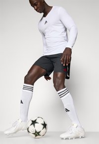 Chemise blanche à manches longues avec logo noir, short noir avec logo rouge, chaussettes blanches à rayures noires, et chaussures de football blanches à côté d'un ballon de football étoilé noir.
