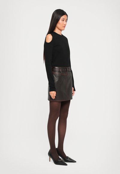 Woman with long dark hair wearing black cold-shoulder top, dark brown leather skirt with belt, black tights, and black pointed-toe heels, standing sideways.