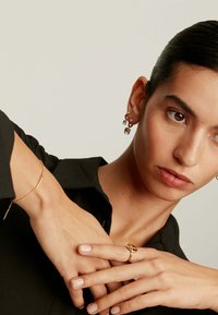 Joyería de oro, que incluye una pulsera, un anillo y pendientes colgantes con piedras transparentes. La modelo lleva una camisa negra, exhibiendo los accesorios.