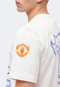 Jeune personne portant une chemise blanche avec un écusson du Manchester United Football Club sur la manche, tournée vers la gauche sur un fond uni.