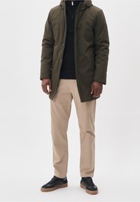 Man som bär en hoodie i olivgrön färg, svart tröja, beige byxor och svarta sneakers med gummisulor, står framför en vit bakgrund.