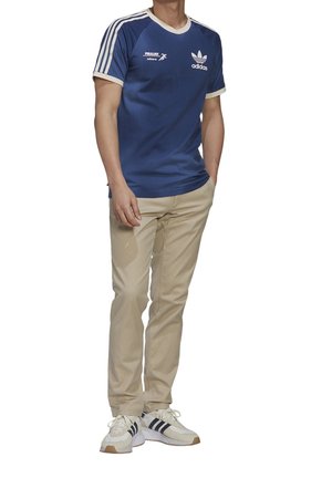 Niebieska koszulka Adidas z białymi wykończeniami i logo, w połączeniu z beżowymi spodniami. Model nosi białe buty sportowe z granatowymi paskami i jasnobrązowymi akcentami.