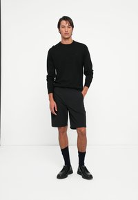 Svart langermet skjorte kombinert med svarte shorts, begge i en glatt stoff. Modellen har på seg svarte sokker og sko med et minimalistisk design.
