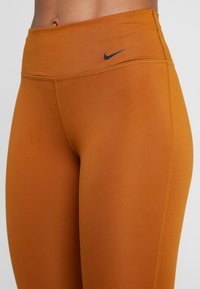 Pomarańczowe leginsy wysokiej talii o gładkiej fakturze, z czarnym logo Nike na pasie i kontrastującym żółtym przeszyciem.
