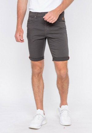 Homme portant un short gris foncé retroussé, des baskets blanches et une chemise blanche sur un fond clair uni.