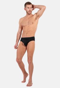 Schwarze Herrenbadehose mit glatter Textur, ausgestattet mit einem unterstützenden Gummibund und einem konturierten Beutel-Design, präsentiert an einem Modell vor einem schlichten Hintergrund.