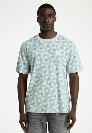 Homme portant une chemise à manches courtes vert clair avec un motif géométrique abstrait vert foncé et un pantalon gris, debout devant un fond blanc.