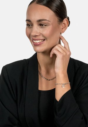 Silver chain necklace with elongated links, paired with a silver bracelet featuring a round charm. Model wears a black top and jacket.