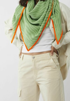 Personne portant un pantalon beige clair, un crop top blanc côtelé, une veste crème et une écharpe à motif vert avec des bords orange vif autour du cou.