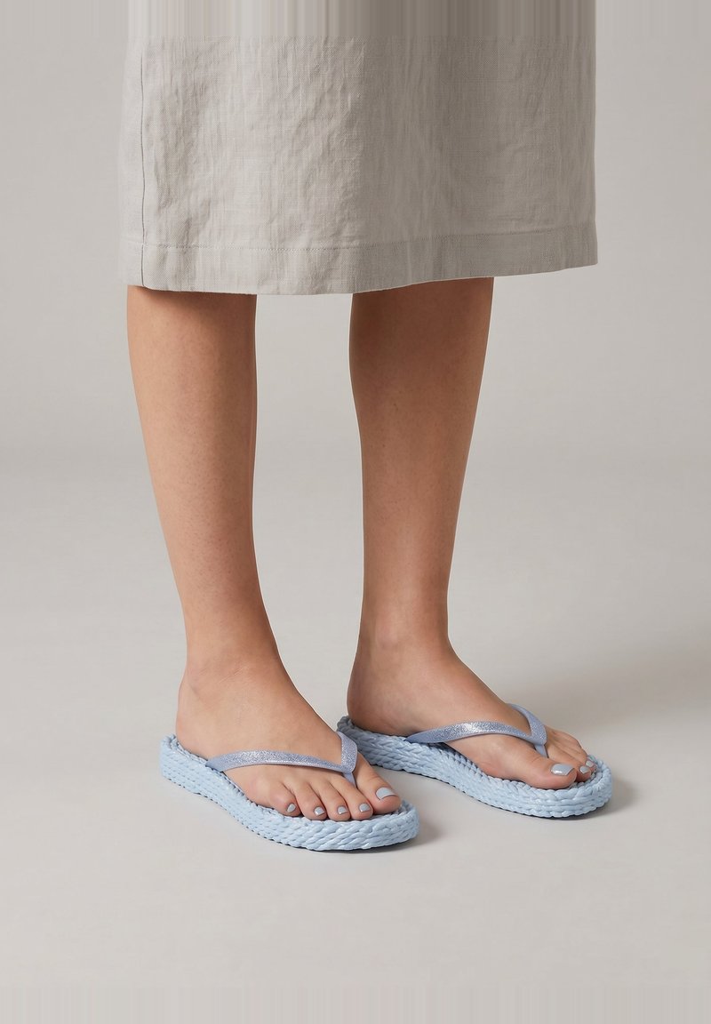 Blote voeten met lichtblauwe gevlochten flip-flop slippers gecombineerd met een beige linnen midi-rok tegen een neutrale achtergrond.