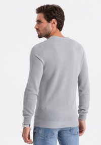 Hellgrauer Strickpullover mit strukturiertem Muster, Rundhalsausschnitt sowie gerippten Bündchen und Saum. Getragen mit blauen Jeans.