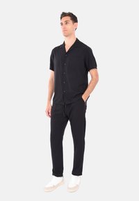Camicia nera a maniche corte con bottoni e tasca, abbinata a pantaloni neri dal taglio morbido e scarpe da ginnastica bianche su uno sfondo chiaro.