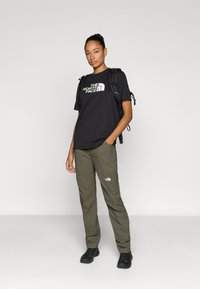 Μαύρο T-shirt με κοντά μανίκια και λευκό λογότυπο, σε συνδυασμό με χακί παντελόνια και μαύρα παπούτσια. Φοράει μαύρο σακίδιο στο ένα του ώμο.