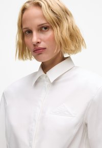 En kvinne med blondt, kort, bølget hår iført en hvit skjorte med buttons og en brettet lommefyrkant foran en ensfarget hvit bakgrunn.