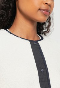 Veste en polaire crème avec un panneau texturé bleu marine ; dotée d'un col rond et d'une fermeture à bouton rond bleu marine à l'avant.