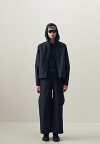 Black hoodie with a front zipper, paired with a black long-sleeve top and wide-legged pants. Accessories include dark sunglasses and black shoes.