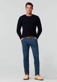 Pull en mailles torsadées bleu marine, jean bleu et chaussures à lacets beige clair avec des lacets marron. L'ensemble présente un design classique et ajusté.