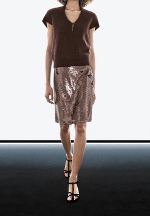 Femme marchant avec un pull marron à manches courtes, une jupe brillante en sequins bronze, des sandales noires à talons hauts et lanières, et un collier délicat.