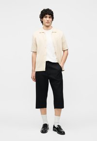 Ένας νεαρός άνδρας στέκεται φορώντας ένα μπεζ κοντομάνικο πουκάμισο με κουμπιά, άσπρο μπλουζάκι, μαύρο φαρδύ κοντό παντελόνι, λευκές κάλτσες και μαύρα μοκασίνια.