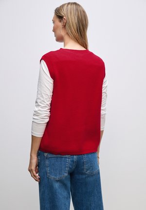 Roter ärmelloser Strickpullover mit gerippter Textur, getragen über einem weißen Langarmshirt. Kombiniert mit blauen weiten Denim-Jeans.