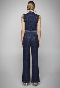 Mono de encaje azul marino con cuerpo ajustado, piernas acampanadas, bordes con forma de concha y un cinturón de tela en la cintura. El detalle en la parte posterior incluye una cremallera.