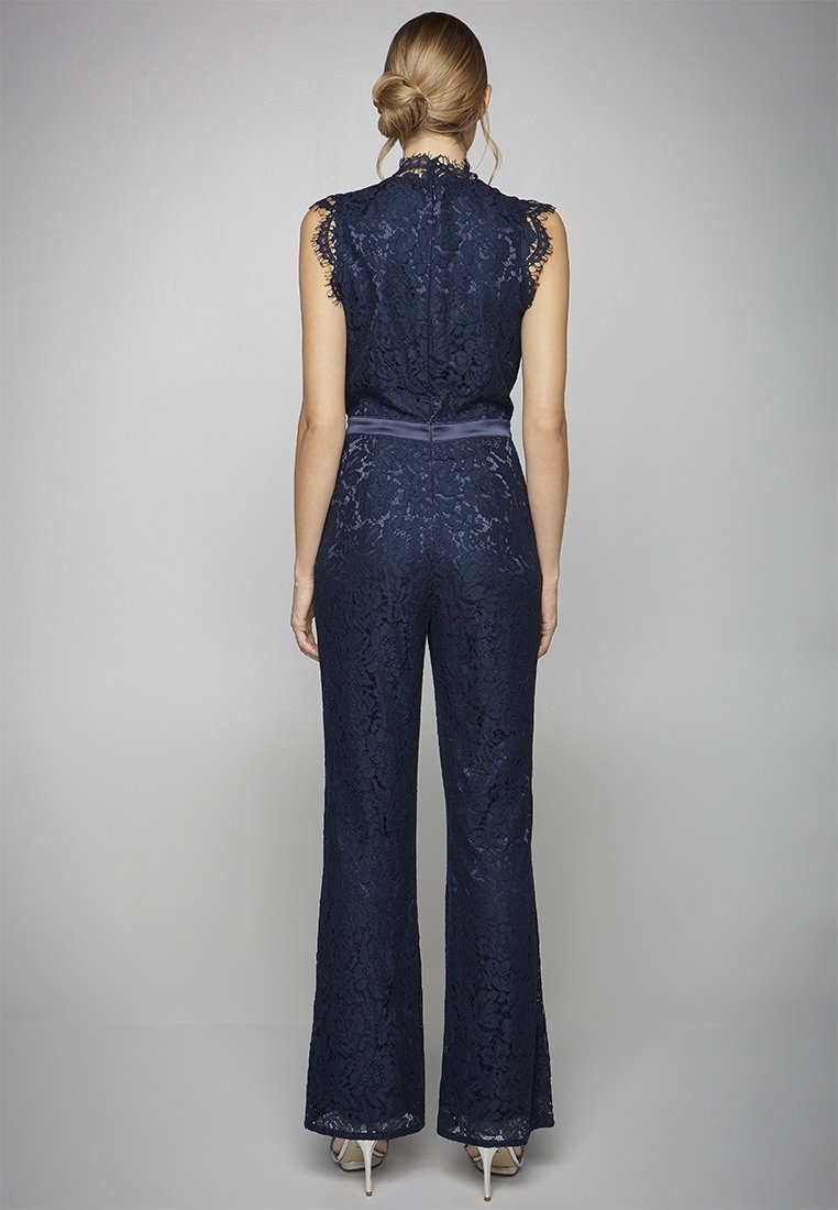 Mono de encaje azul marino con cuerpo ajustado, piernas acampanadas, bordes con forma de concha y un cinturón de tela en la cintura. El detalle en la parte posterior incluye una cremallera.