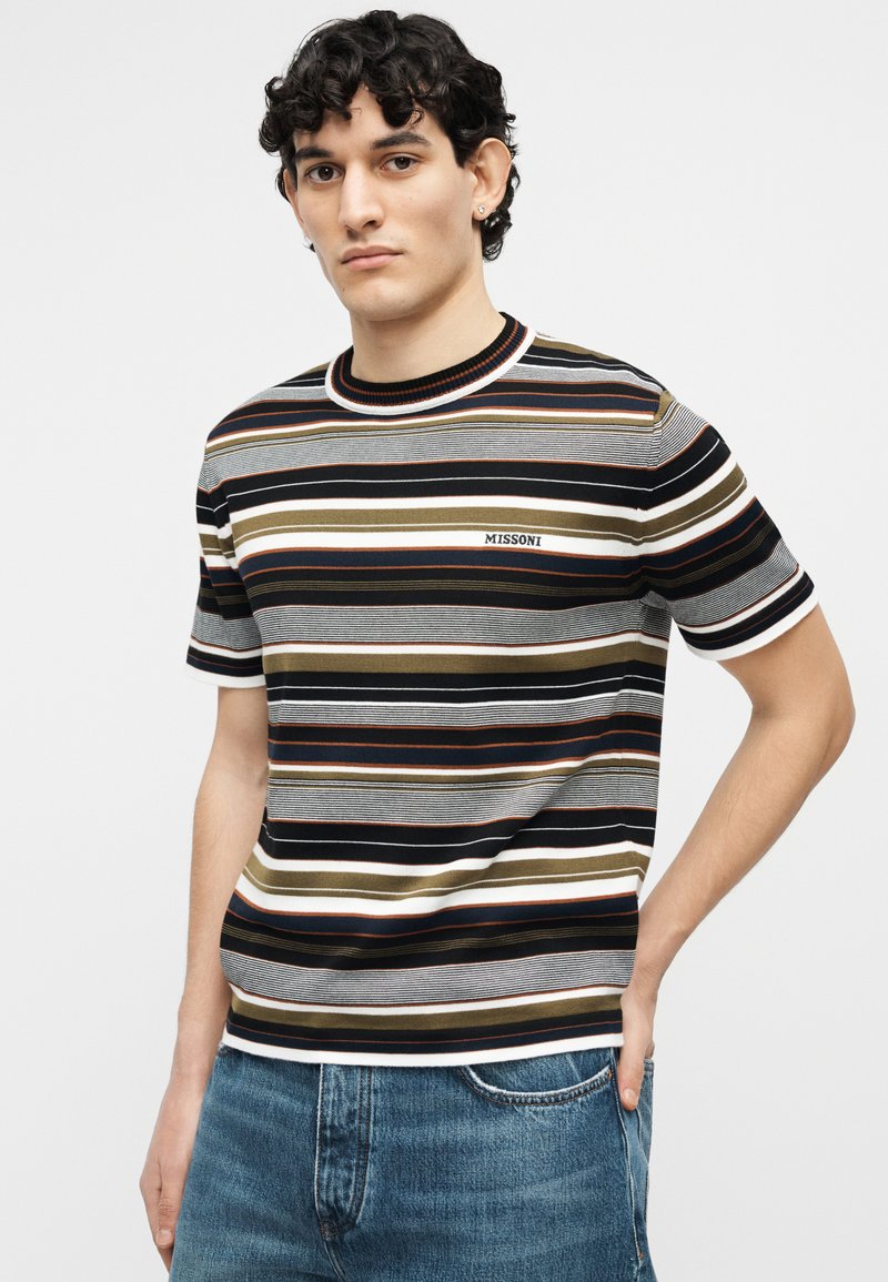 Young man wearing a striped short-sleeve shirt with "MISSONI" logo on the chest and blue denim jeans, standing with one hand in pocket.