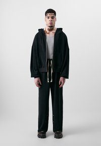 Black zip-up hoodie with two front pockets, paired with loose black pants featuring drawstrings and side snap accents. Textured fabric and athletic shoes.