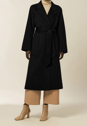 Femme portant un long manteau noir noué à la taille, un pantalon large beige, des bottines beiges et un pull à col roulé beige.