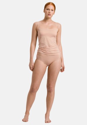 Donna con capelli rossi che indossa una canottiera e slip color carne, in piedi a piedi nudi davanti a uno sfondo chiaro e uniforme.