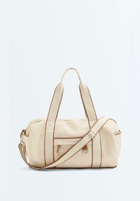 Sac fourre-tout en toile beige avec garniture marron, double poignée, bandoulière réglable, poche avant zippée et anneau en D métallique sur fond uni.