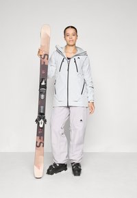 Lyžiarsky outfit s ľahko šedou nepremokavou bundou s čiernymi akcentmi, zodpovedajúcimi ľahko šedými nohavicami a párom hnedo-čiernych lyží.