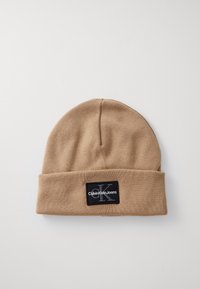 Beige Strickmütze mit umgeschlagenem Rand, versehen mit einem schwarzen rechteckigen Patch mit weißem Textlogo. Weicher Stoff und einfaches Design.