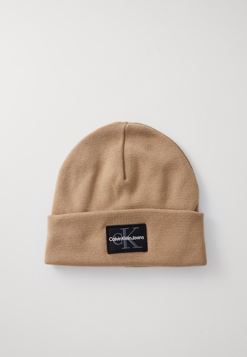 Beige Strickmütze mit umgeschlagenem Rand, versehen mit einem schwarzen rechteckigen Patch mit weißem Textlogo. Weicher Stoff und einfaches Design.