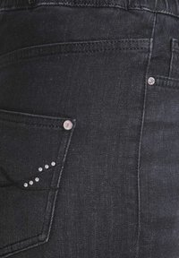 Zwarte denim jeans met een achterzak voorzien van zilverkleurige studs en een glanzend accent. De stof heeft een textuur en is licht reflecterend.