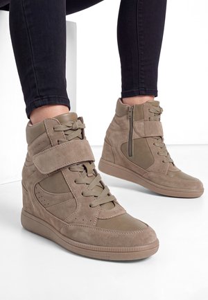 Taupefarbene Keil-Sneakers mit Schnürsenkeln, Klettverschluss und seitlichem Reißverschluss, getragen mit schwarzen, eng anliegenden Hosen vor weißem Hintergrund.