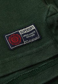 Groene gestructureerde stof met een marineblauw etiket waarop "Superdry Vintage Athletic Outfitters" staat en een rood rond logo.