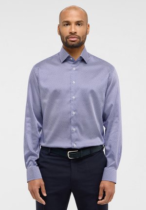 Camicetta a maniche lunghe di colore viola con un motivo geometrico, chiusura a bottoni e colletto alla francese, abbinata a pantaloni scuri e una cintura nera.