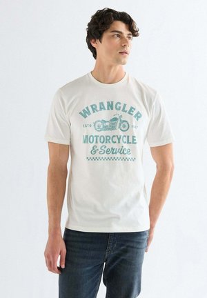 Junger Mann trägt ein weißes Wrangler T-Shirt mit einem blauen Motorradmotiv, kombiniert mit dunkelblauen Jeans, steht vor weißem Hintergrund.