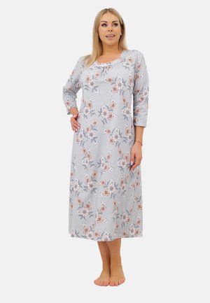 Kobieta stoi boso, ubrana w jasnoszary kwiecisty, długi rękawony nocny strój z różowymi i białymi kwiatami na jednolitym białym tle.