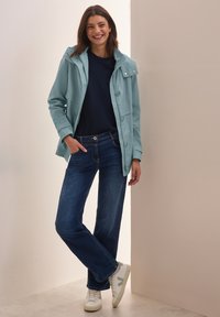 Ljus teal regnjacka med knappar och huva, buren över en marinblå skjorta, i kombination med mörkblå jeans och vita sneakers med gröna detaljer.