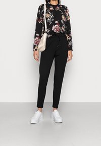 Top nero a maniche lunghe con motivo floreale, colletto rotondo, abbinato a pantaloni neri a sigaretta e sneakers bianche con zeppa, mentre tiene una borsa beige chiaro.